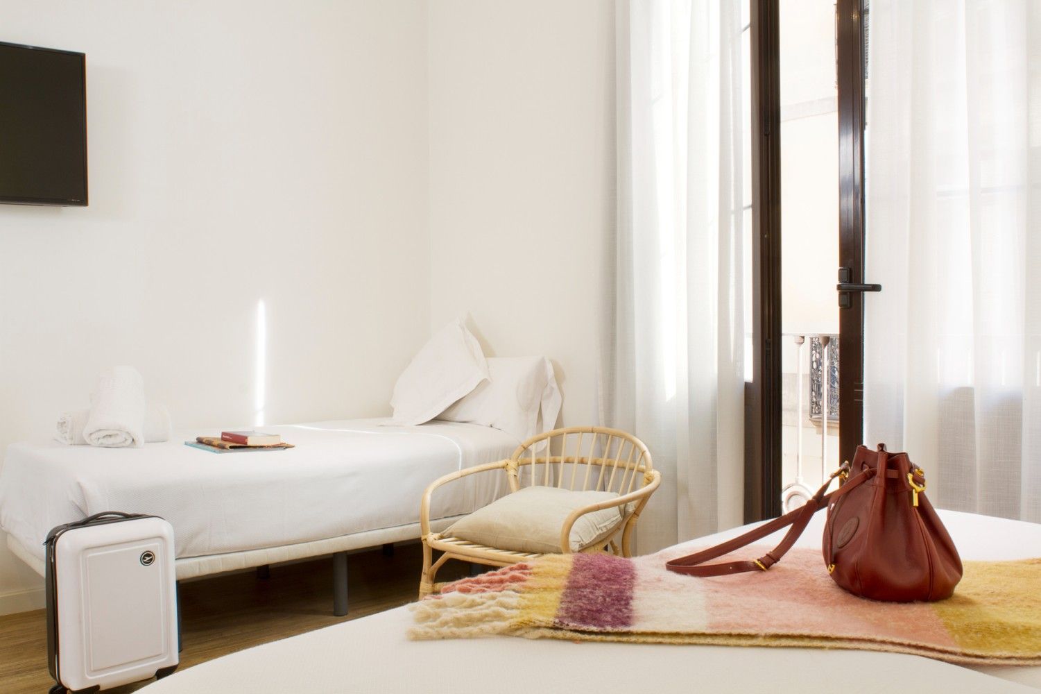 Un dormitorio minimalista y soleado con una cama individual, un sillón de ratán, una maleta blanca.