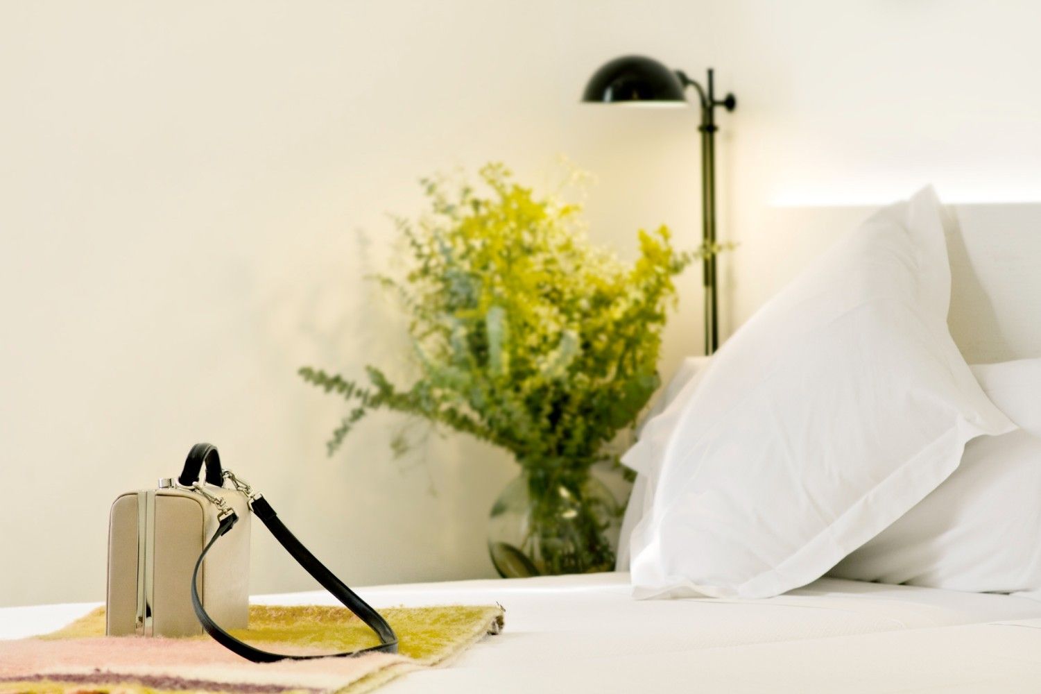 Un pequeño bolso beige reposa sobre una cama junto a un jarrón de flores amarillas.