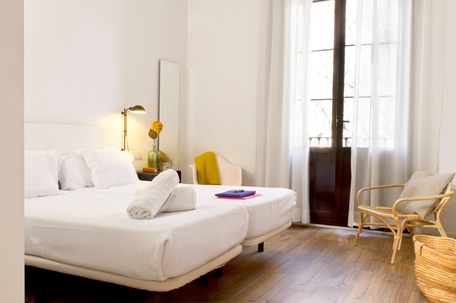 Una habitación de hotel minimalista e iluminada por el sol, con dos camas blancas, una silla de mimbre.