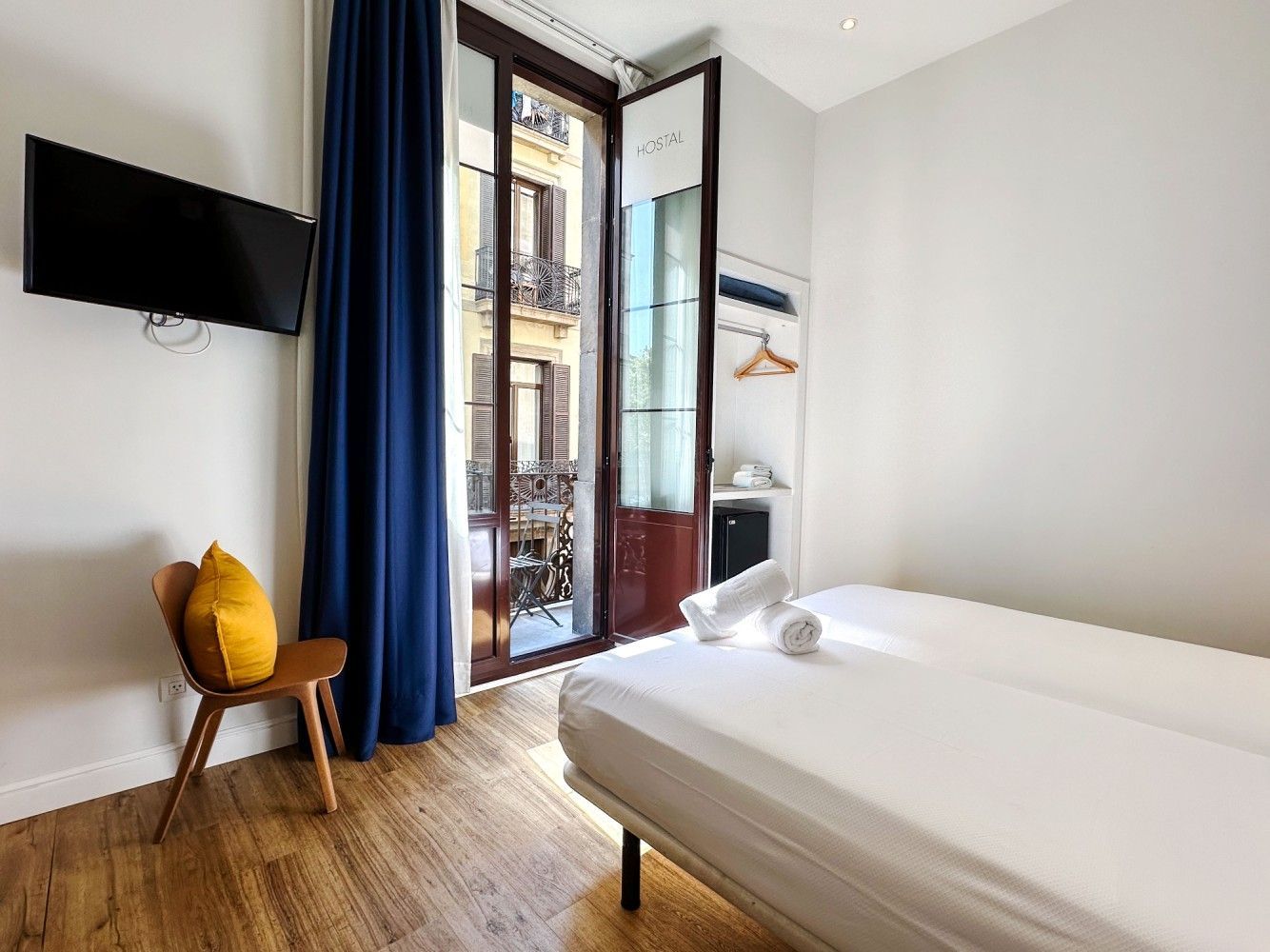 Habitación de hotel luminosa con cama, televisor de pared, cortinas azules y puertas que dan al balcón.