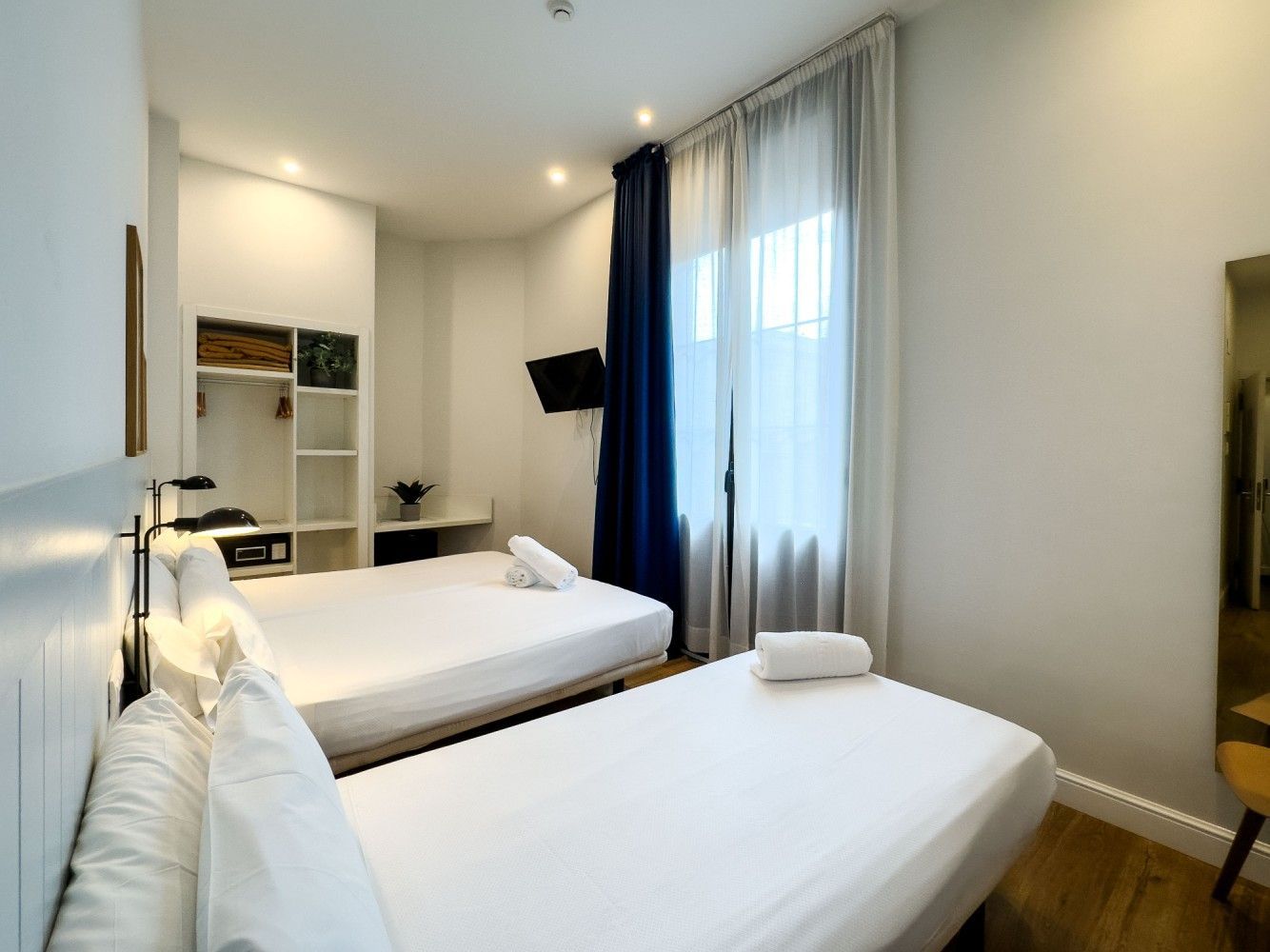 Una habitación de hotel con dos camas blancas, paredes blancas, un mueble de almacenamiento.