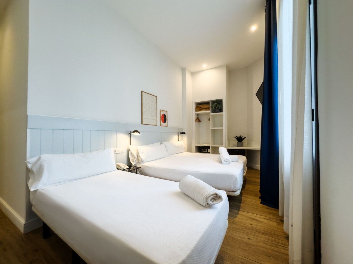 Una habitación de hotel bien iluminada con dos camas blancas, pisos de madera, cortinas azules.