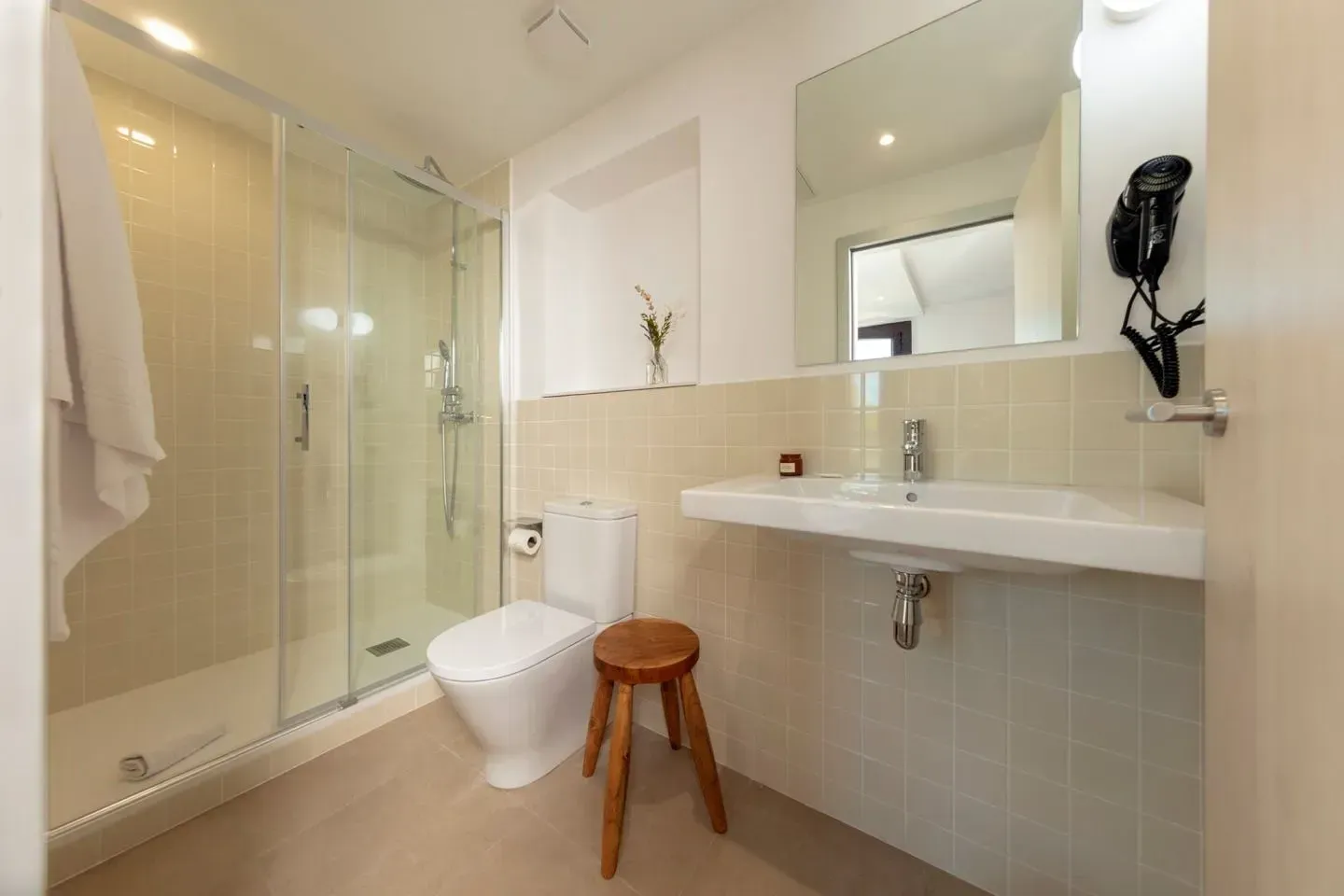 Un baño moderno y luminoso con ducha de cristal, inodoro blanco, lavabo de pared y un taburete de madera.