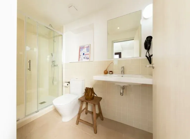 Un moderno baño beige con una ducha de cristal, un inodoro blanco, un lavabo flotante y un pequeño taburete de madera.