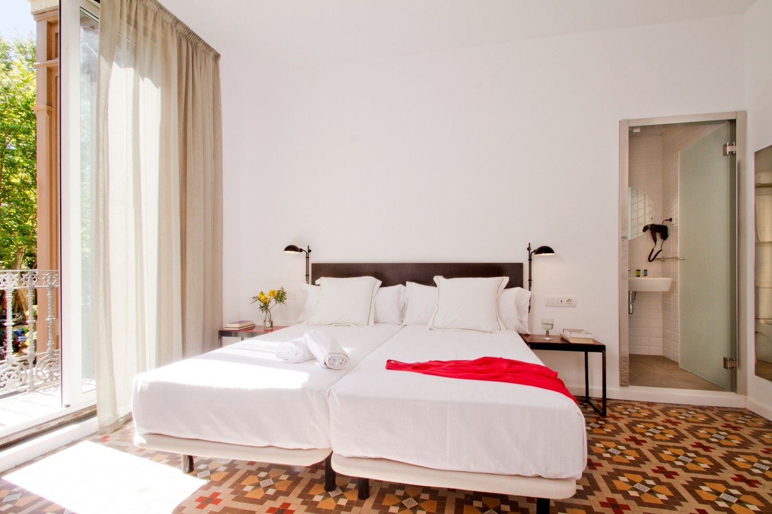 Una habitación de hotel luminosa con dos camas individuales, una alfombra roja, suelos de baldosas estampadas.