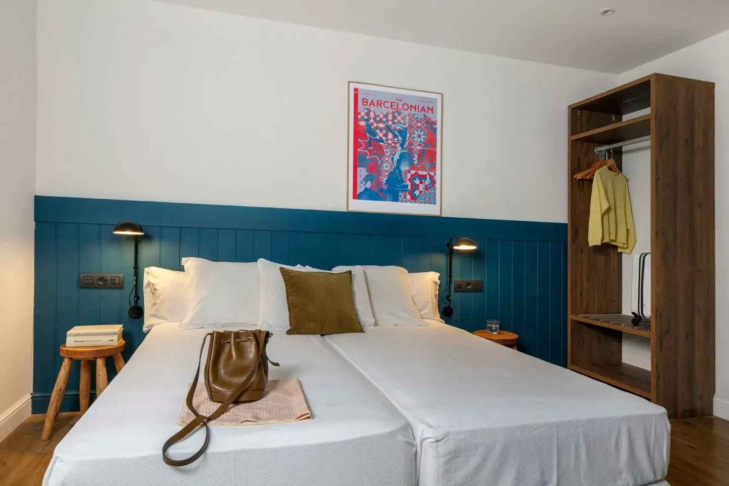 Un dormitorio con dos camas individuales, una cabecera con paneles de color azul verdoso, un taburete de madera.