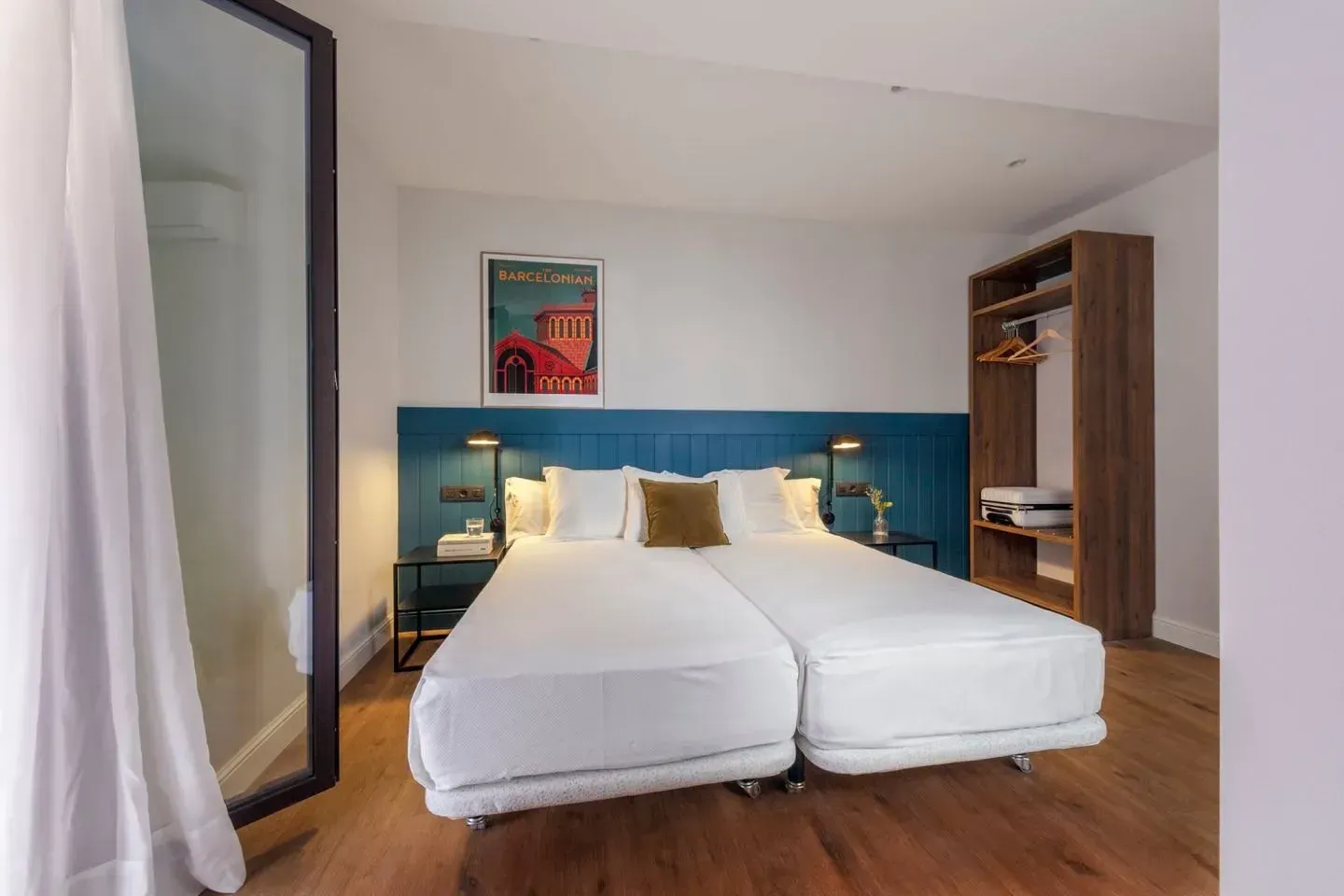 Una habitación de hotel con dos camas individuales juntas, un cabecero azul, un póster vintage y un armario de madera abierto