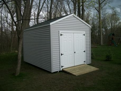 A gray storage shed with white double doors and a wooden ramp. Set in a grassy yard, surrounded by trees.