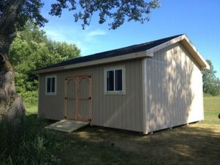 Tan shed with black roof, two windows, and ramp outdoors, set against trees and sky.
