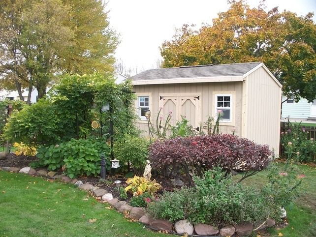 Tan shed with double doors, set in a garden with colorful plants and trees.