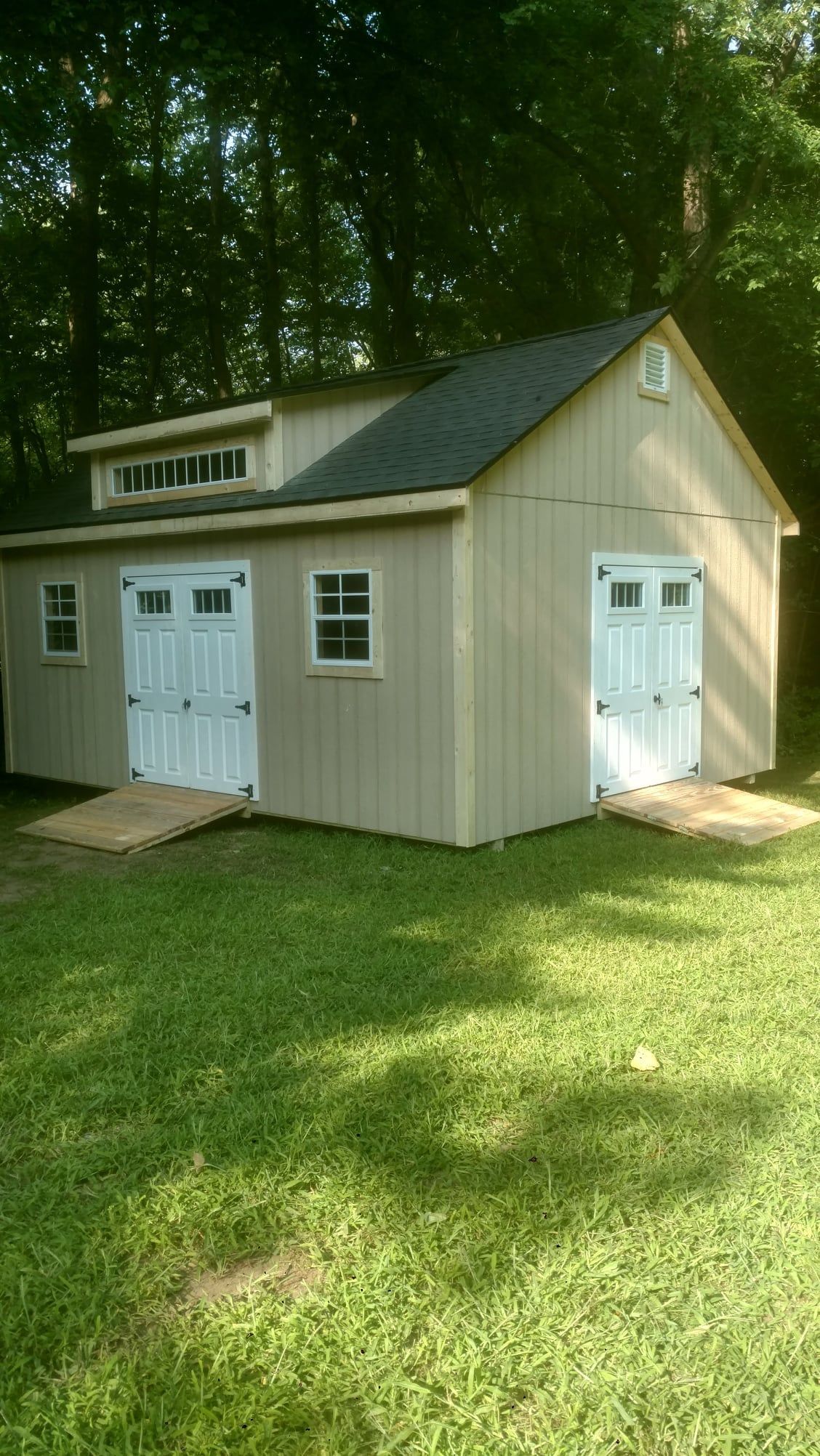Tan shed with white doors, windows, and ramps on green grass, surrounded by trees.