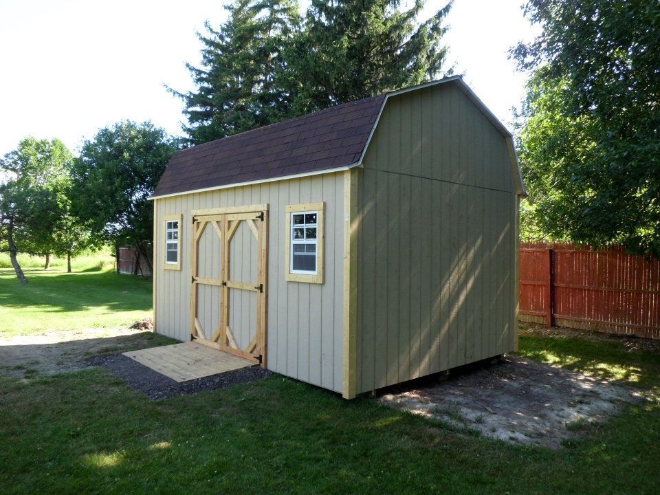 Tan and green barn-style shed with wooden doors and a ramp, set in a grassy yard.