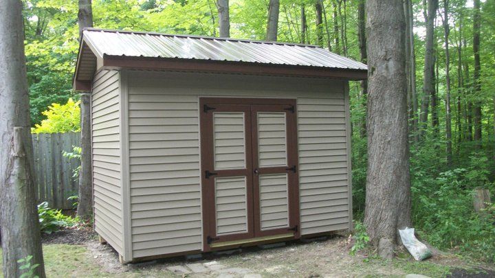 Tan shed with brown trim and metal roof in a wooded area. Double doors centered on the front.