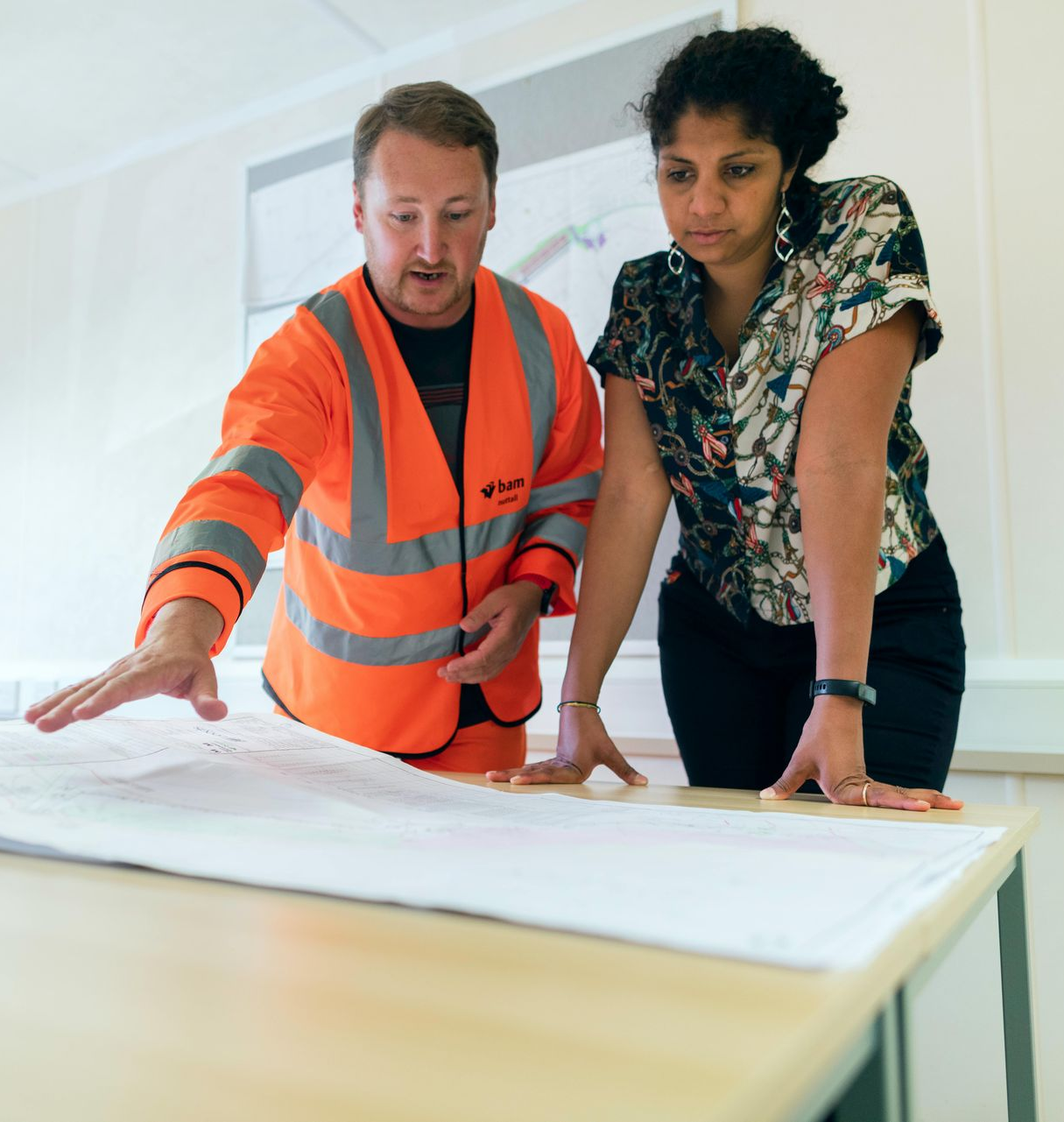 Man in orange vest points at blueprint as woman looks on in office.