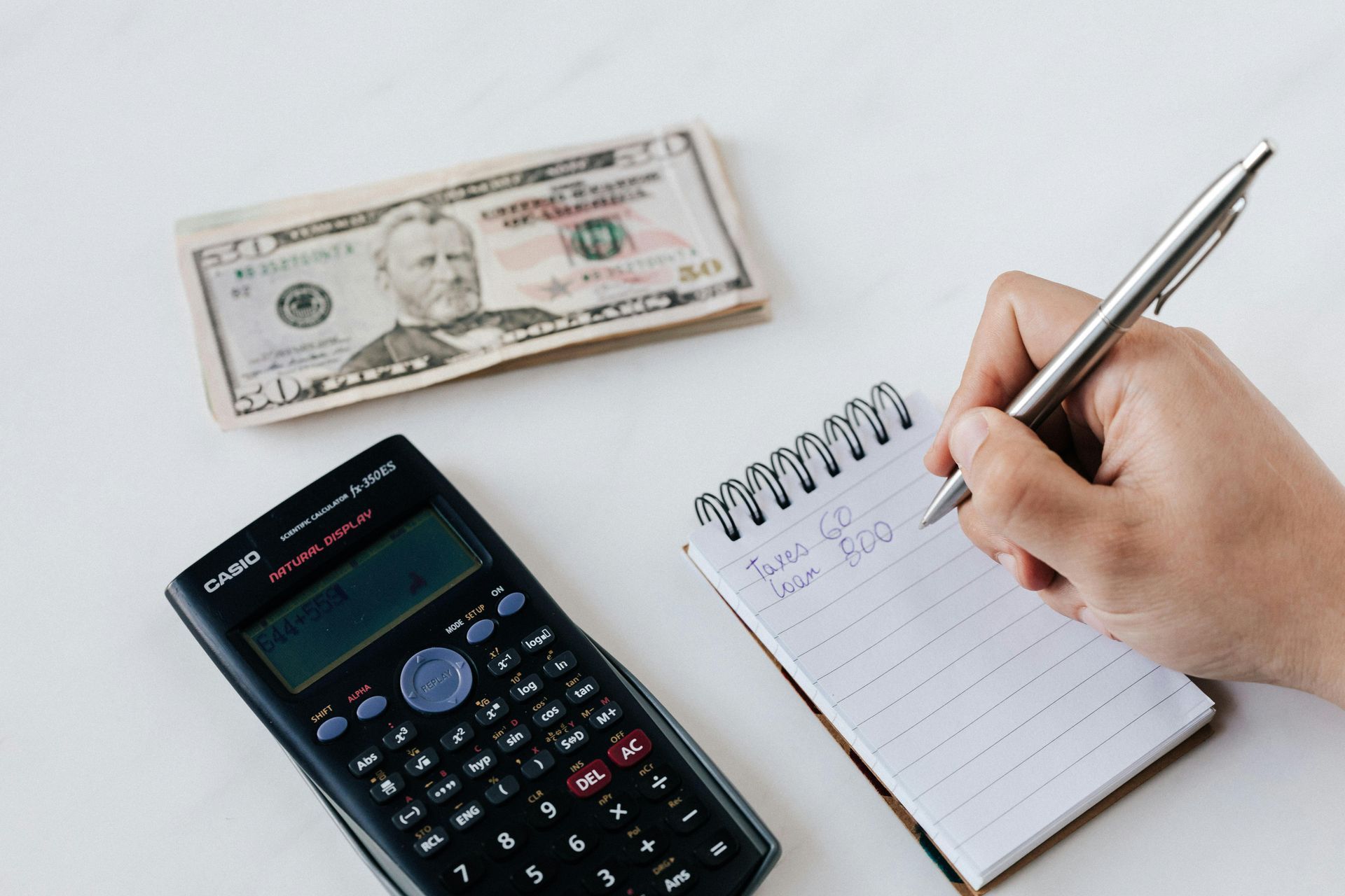 Hand writing in notebook with calculator and cash on a white table.