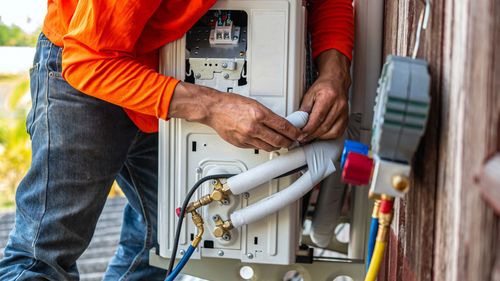 HVAC technician in orange shirt installs an air conditioning unit outside, connecting tubes and hoses.