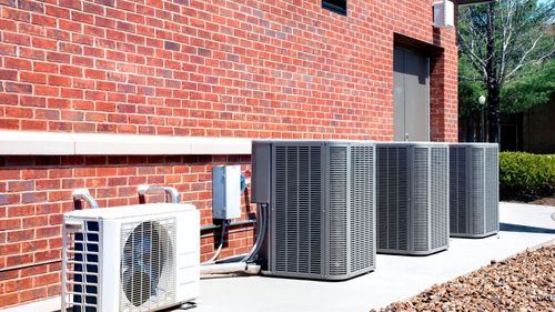 Air conditioning units aligned along a red brick building's exterior wall on a sunny day.
