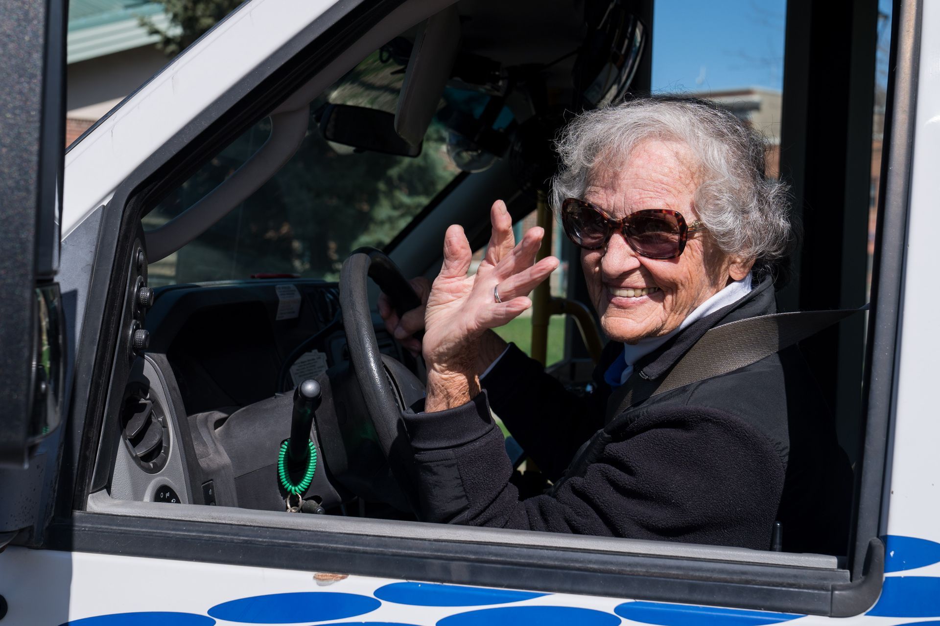 Smiling person in sunglasses waving from the driver’s seat of a white vehicle