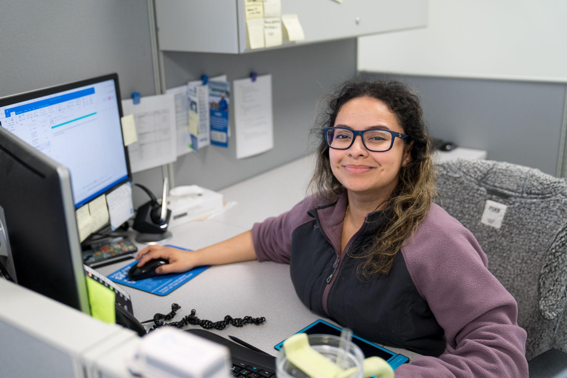 Person smiling at a desk, working on a computer in an office cubicle.