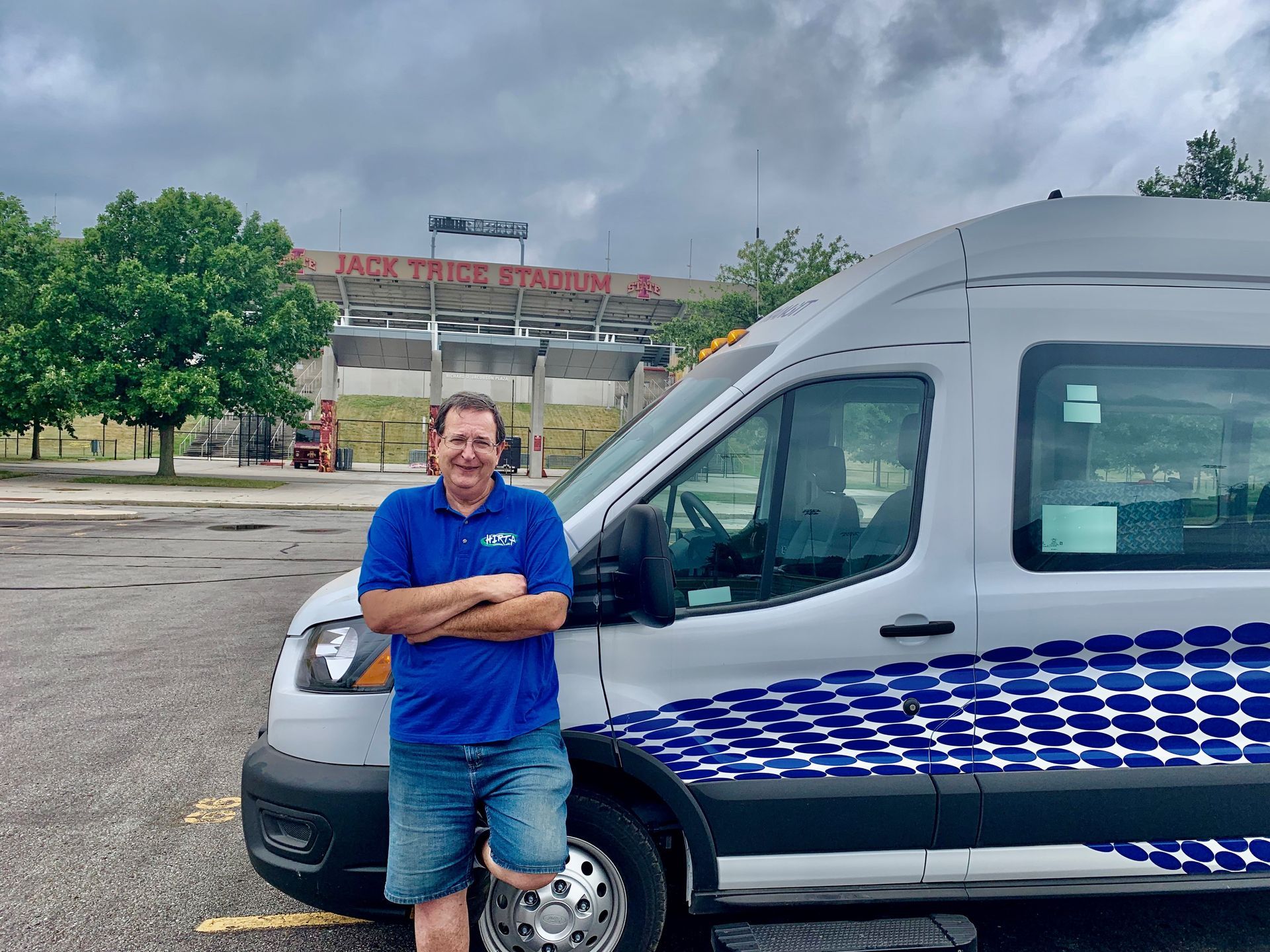 Man in blue shirt leaning on silver van with blue stripe, parked outside a stadium sign.