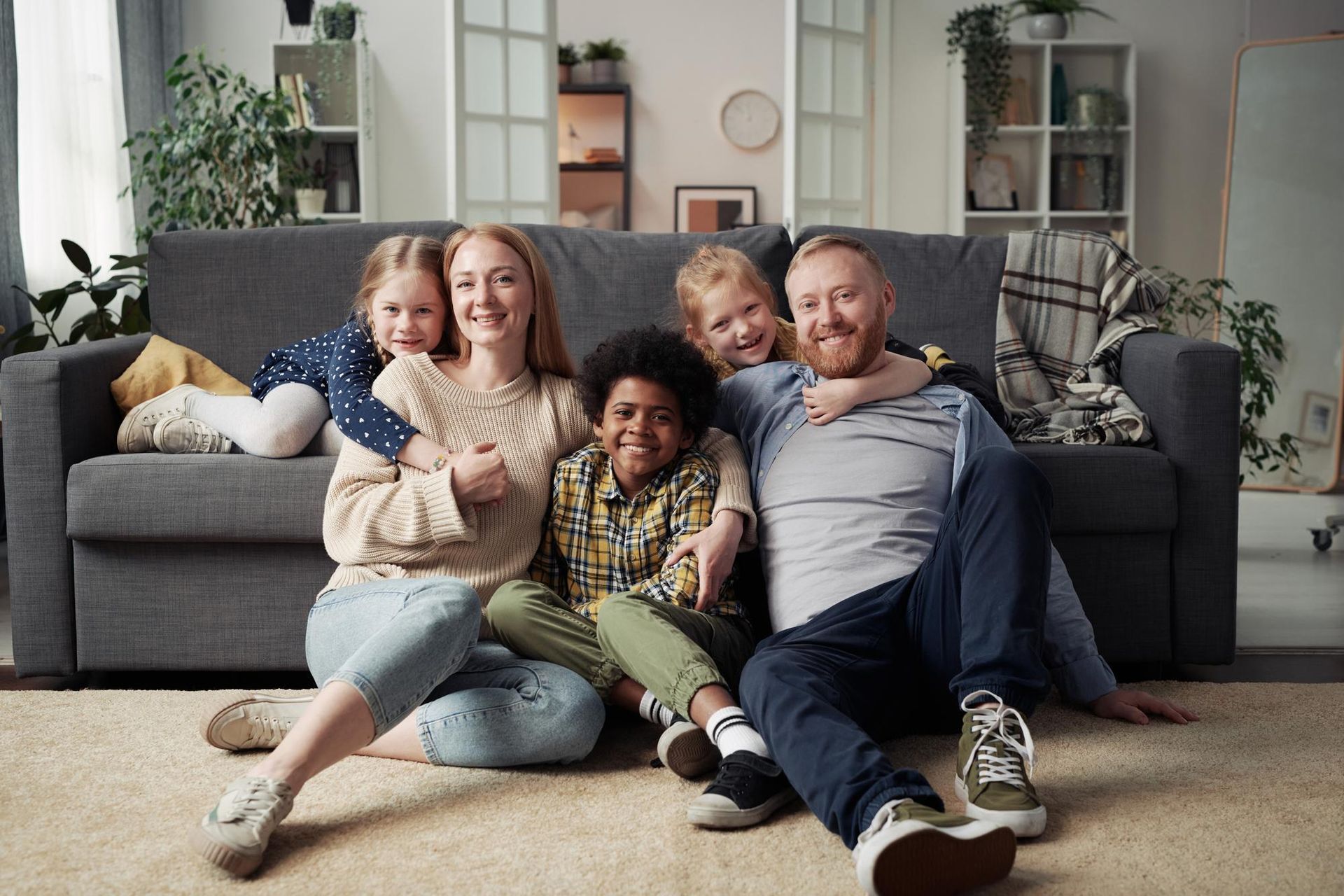 Family of five, diverse and smiling, posing in front of a gray couch in their living room.