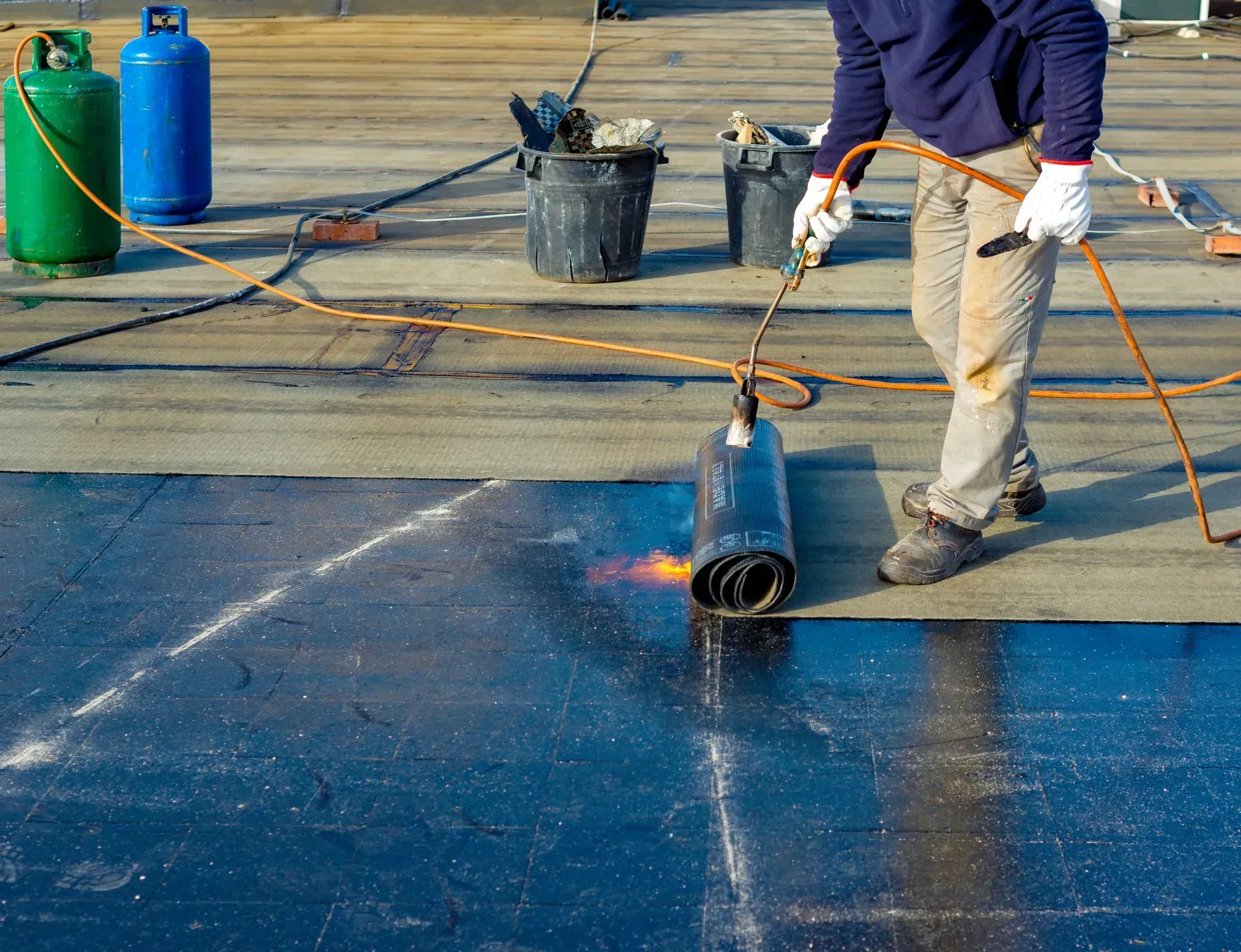 Worker torching a roof membrane, with blue flames on a flat rooftop and safety gas cylinders nearby