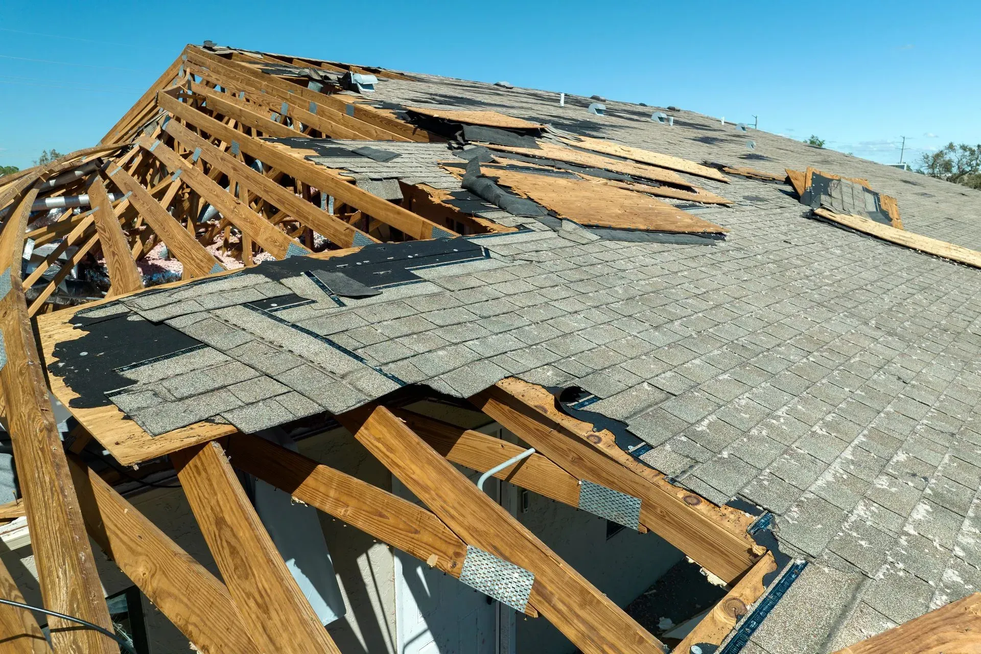 Storm-damaged roof with torn shingles and exposed wooden rafters under a clear sky