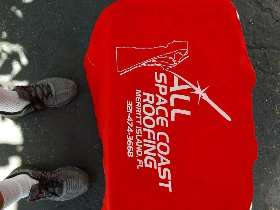 Red beach bag with “Space Coast Running” logo beside gray sneakers on pavement.
