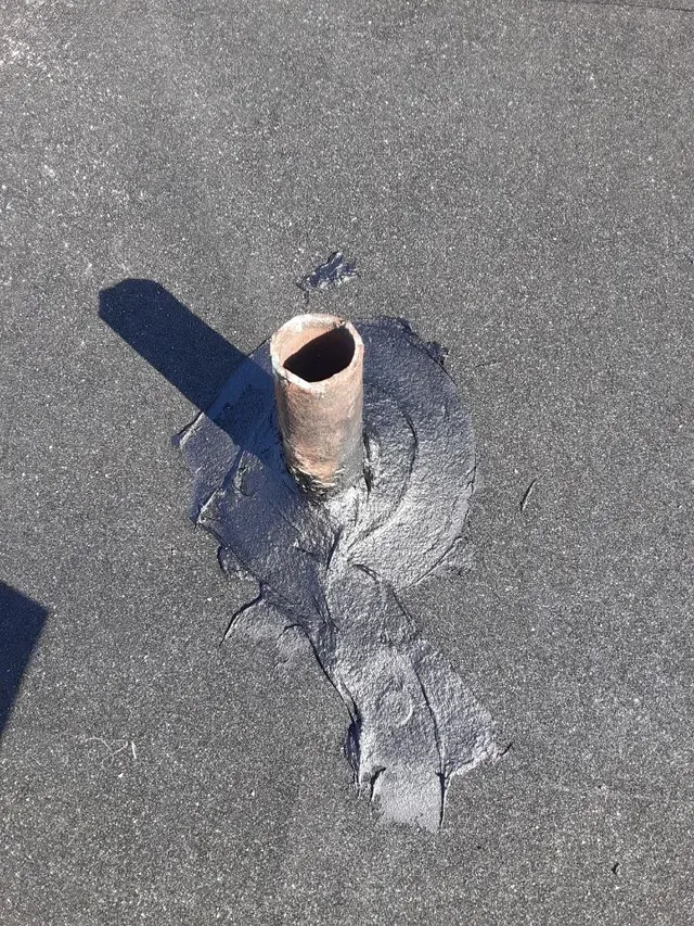 A metal pipe protruding from a flat gray roof with patched roofing material around it.