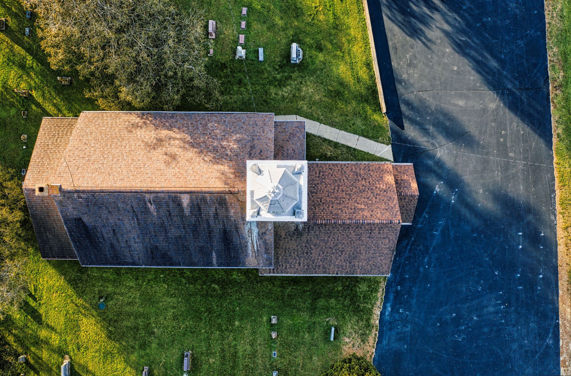Aerial view of a small stone building with a shingled roof beside grass and a paved road.