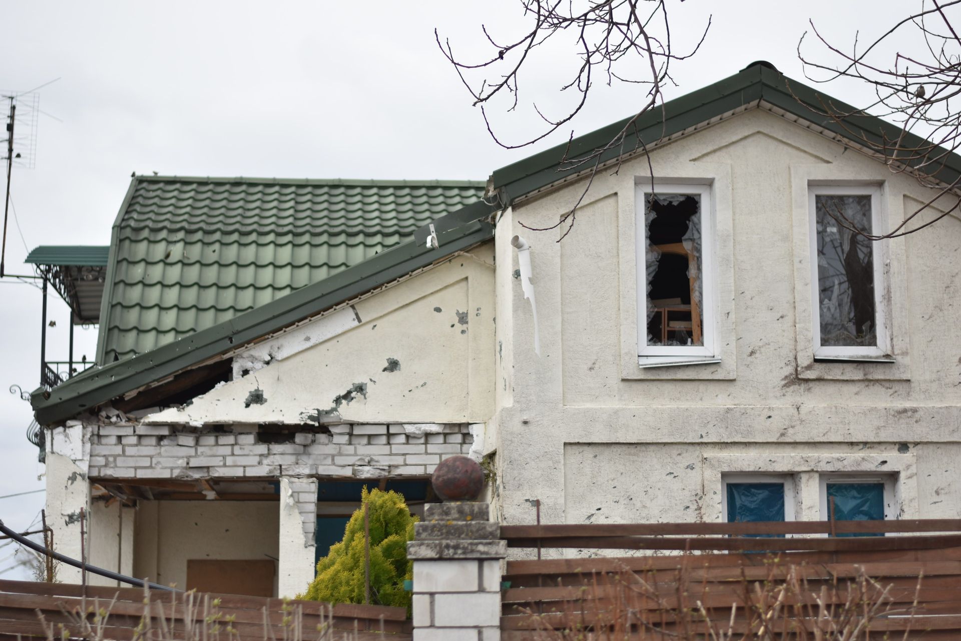 Damaged white house with green roof and broken upper windows behind a fence