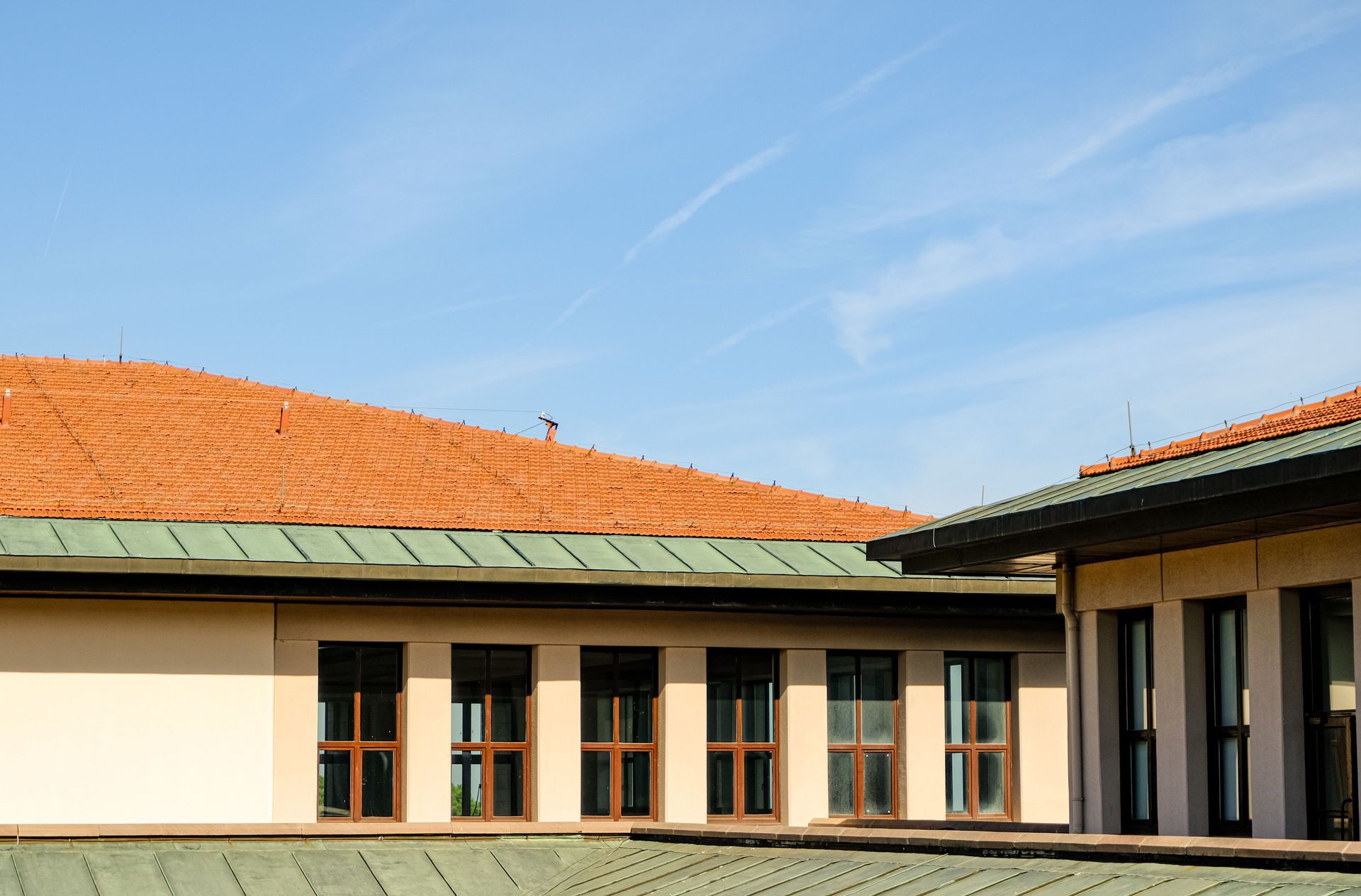 Sunlit building with red-tiled roof, green metal trim, and tall windows under a blue sky