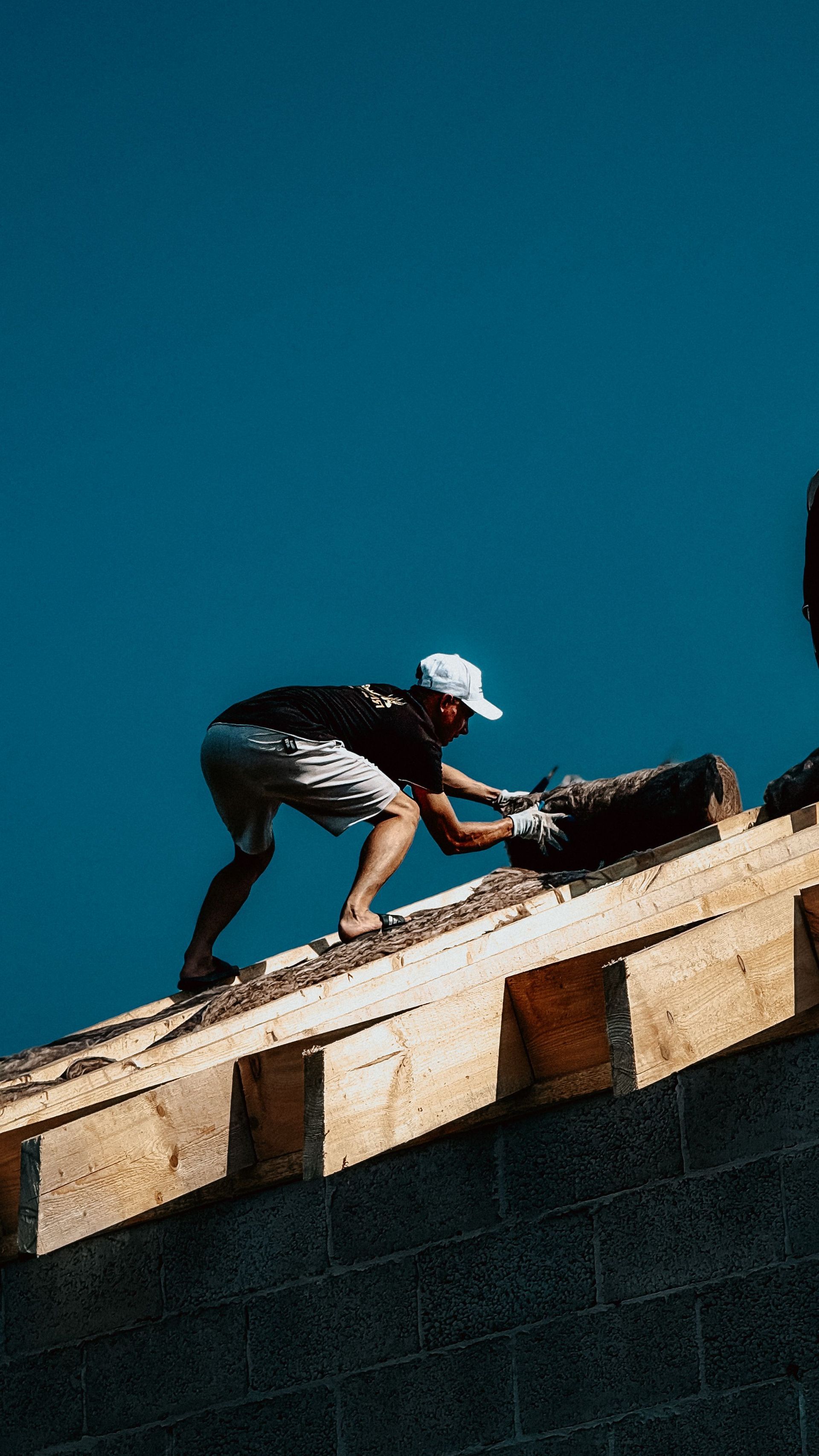 Worker crouching on a sloped rooftop, using tools against a bright blue sky
