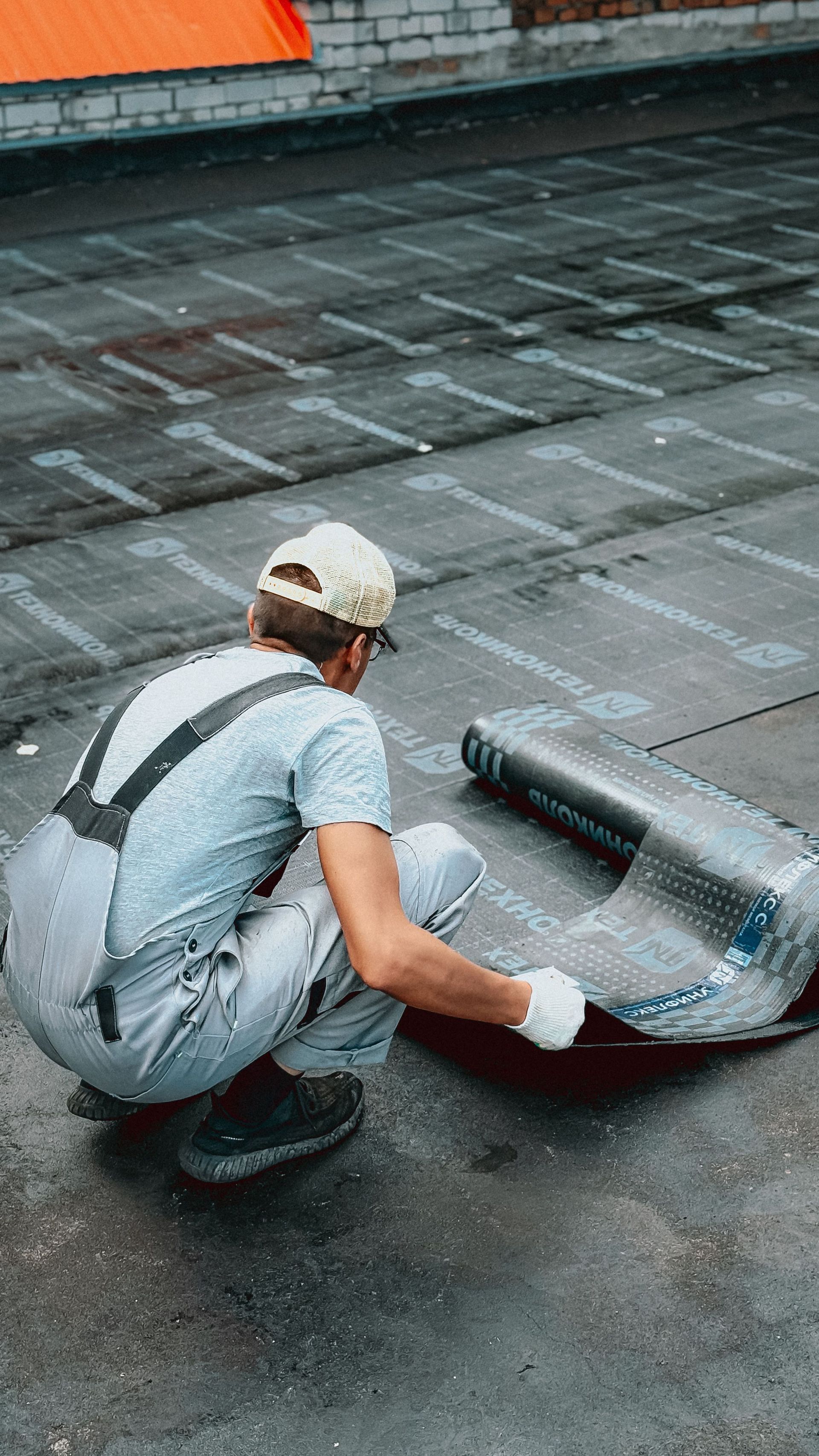Worker unrolling roofing membrane on a flat roof, with a torch roll in hand.