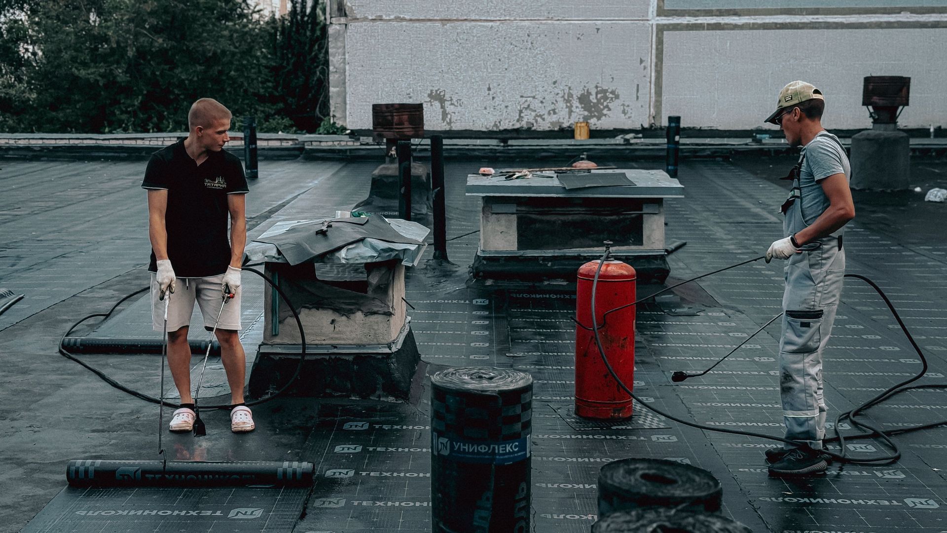 Two workers on a rooftop with equipment, hoses, and a red cylinder on a wet surface.