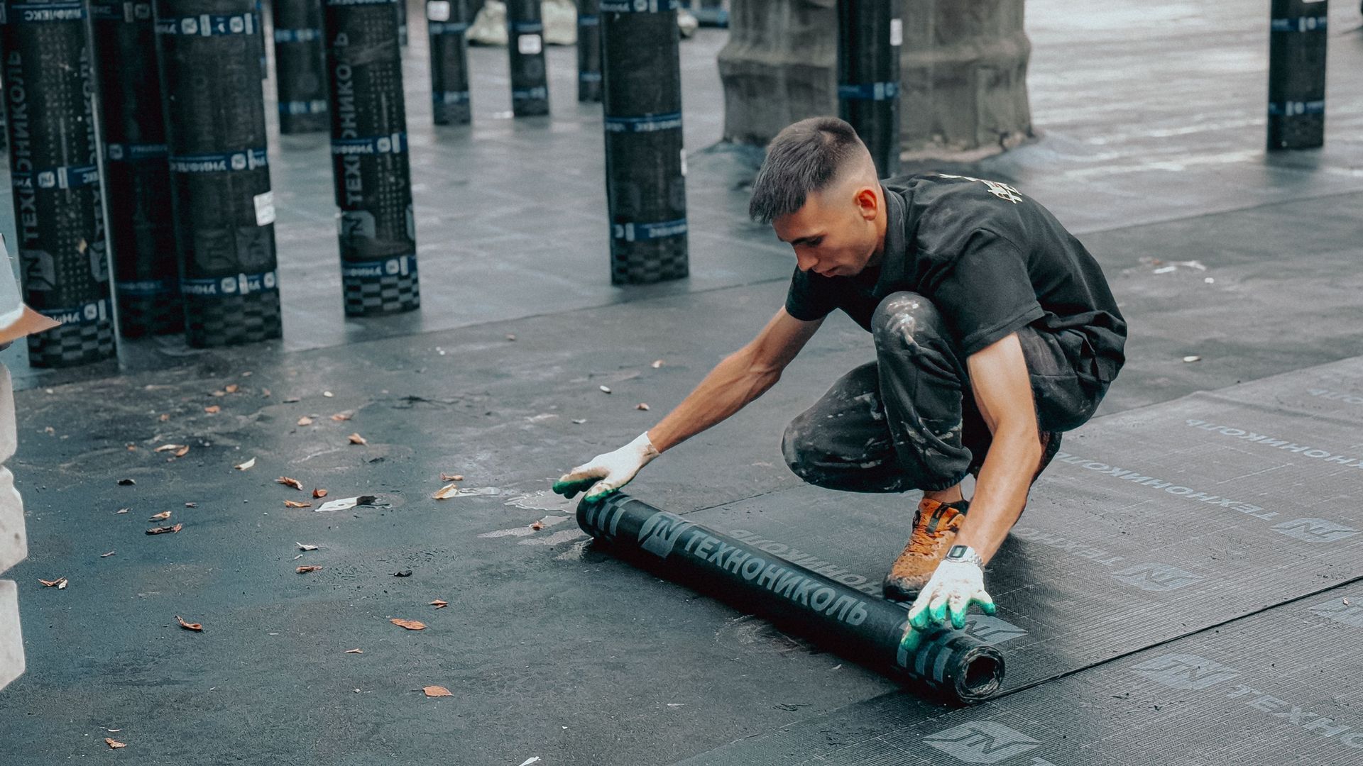 Man crouching on a wet sidewalk, rolling up a dark mat near a row of black posts.
