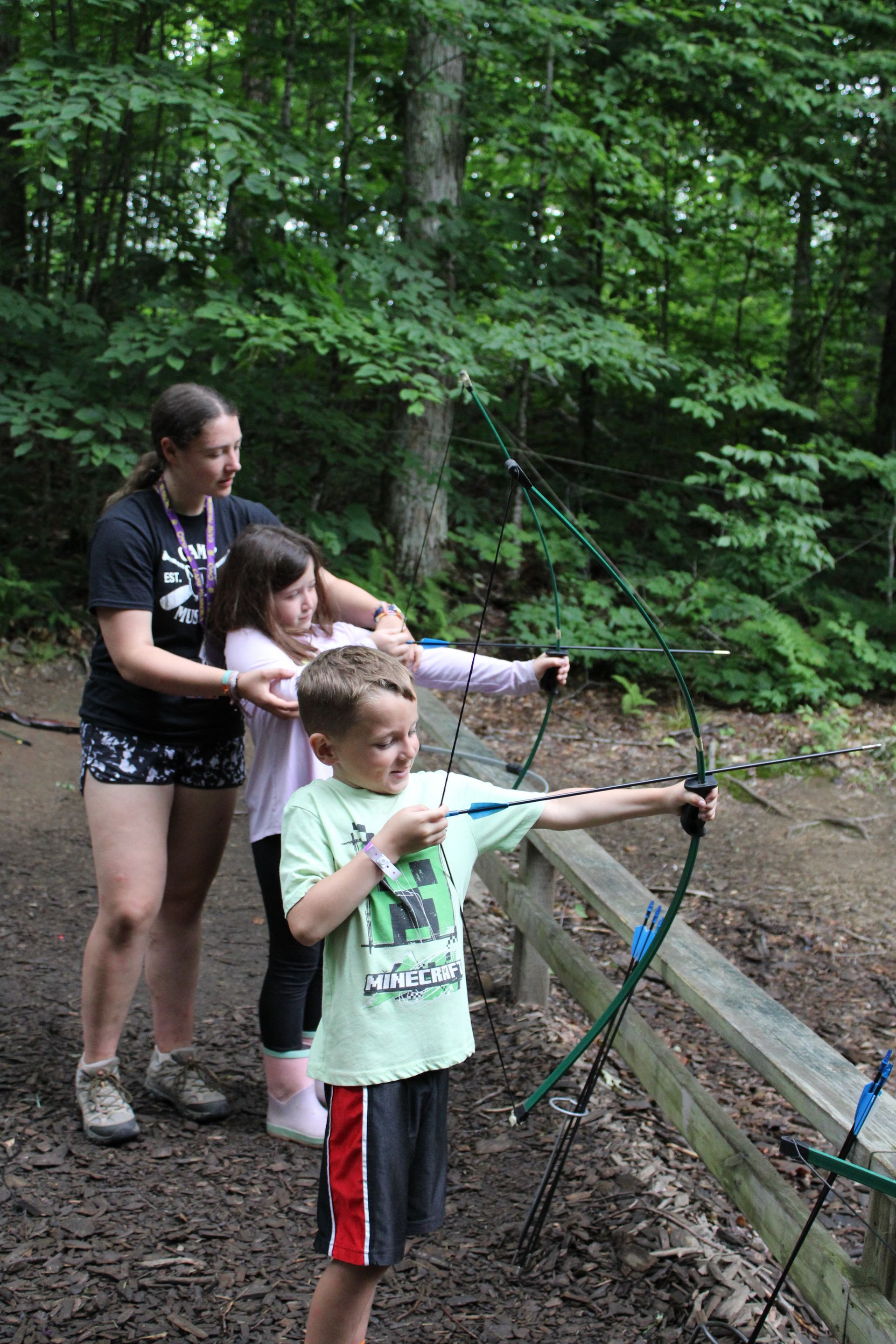 Instructor helps two children with archery in a wooded area. They hold bows, aiming at targets.