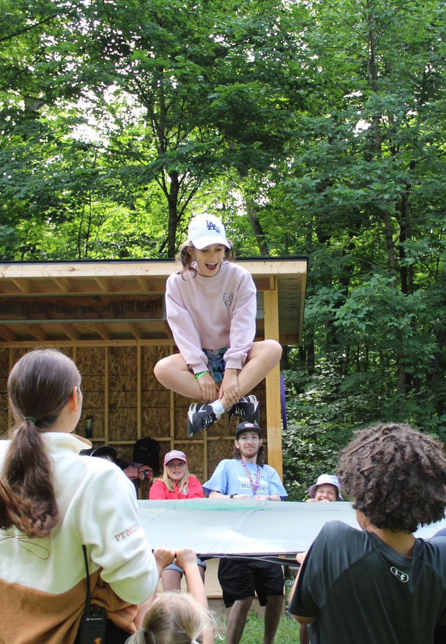 Girl jumps over a board held by children. Other kids watch near a wooden structure outdoors.
