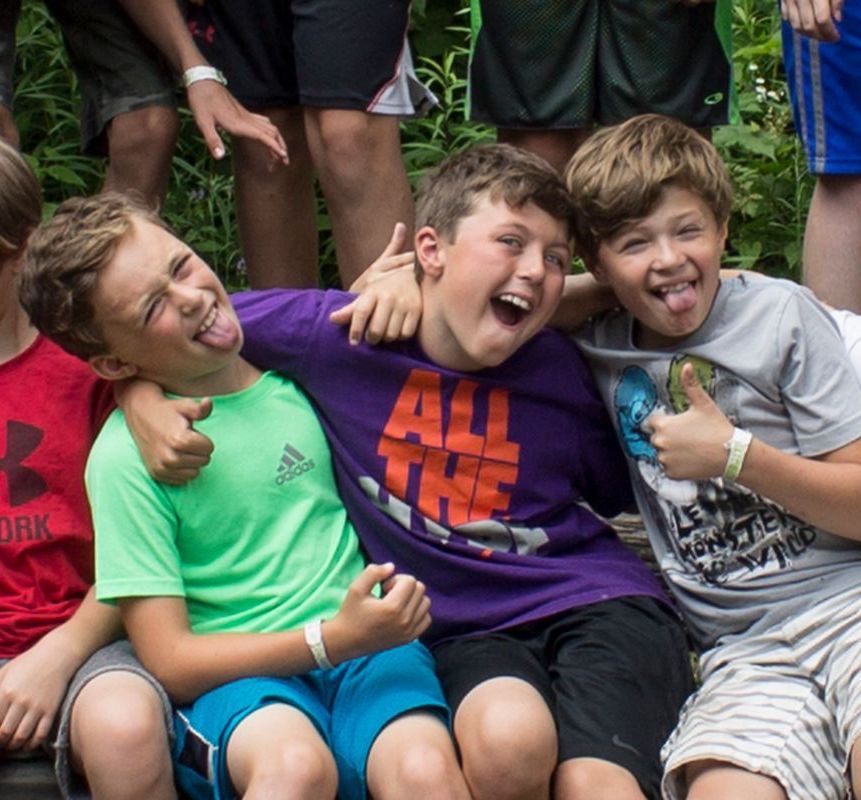 Three boys grinning, sticking out tongues. One in green, one in purple, one in gray. All have arms around each other.