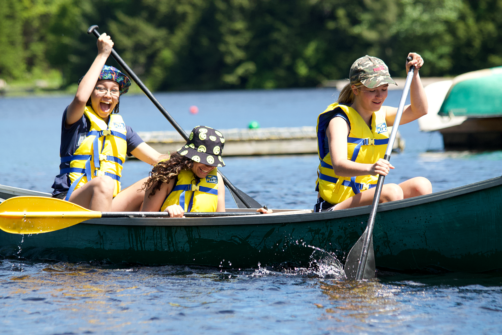 Three people canoeing on a lake, paddling. One person is wearing a polka-dot hat. All are wearing life vests.