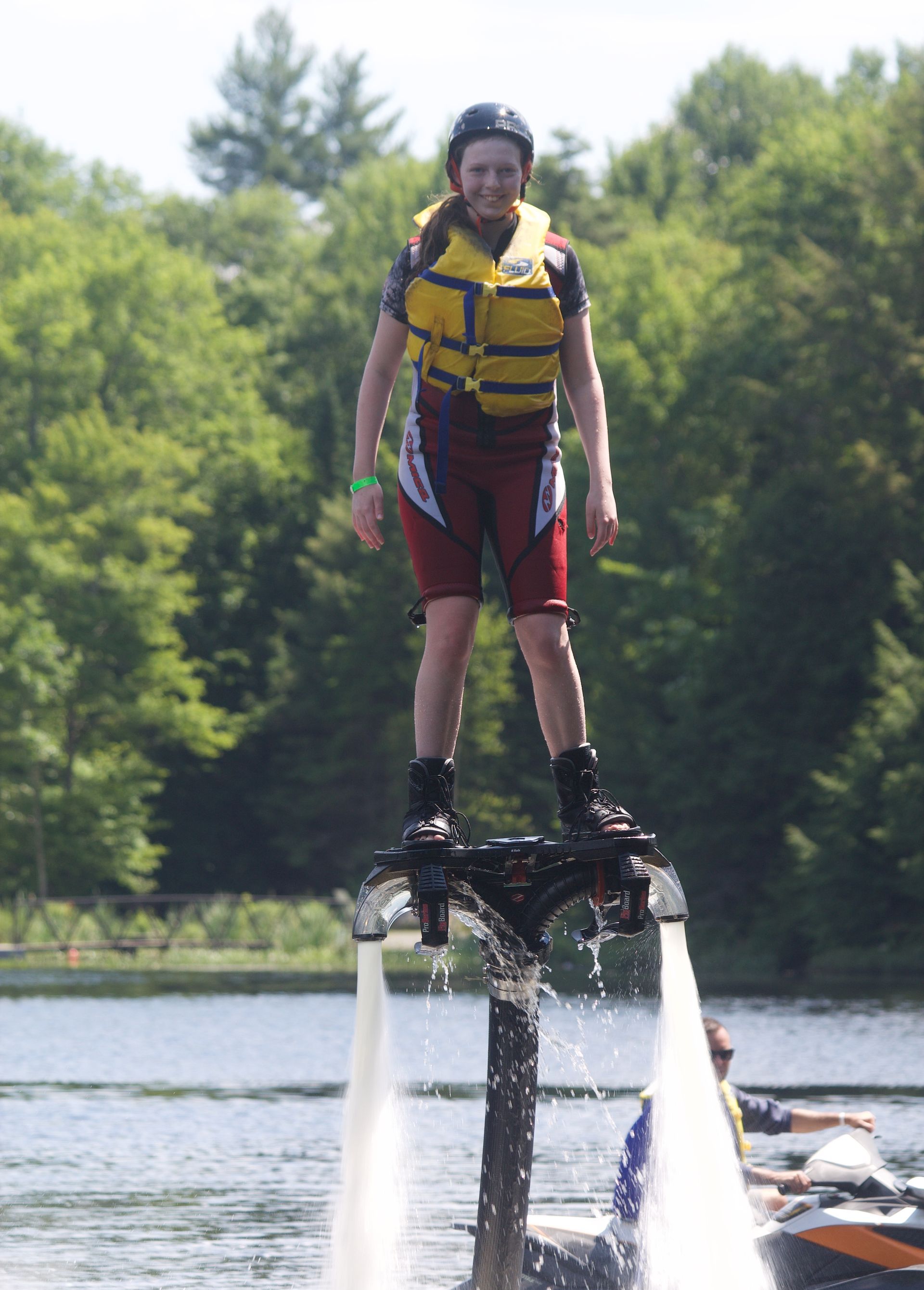 Person on a flyboard, water jets propelling them upward. Wearing a helmet and life vest, standing over water.