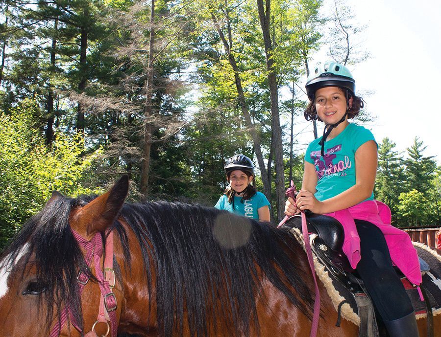 Two girls on horseback, smiling. One in blue shirt and helmet, forest background.