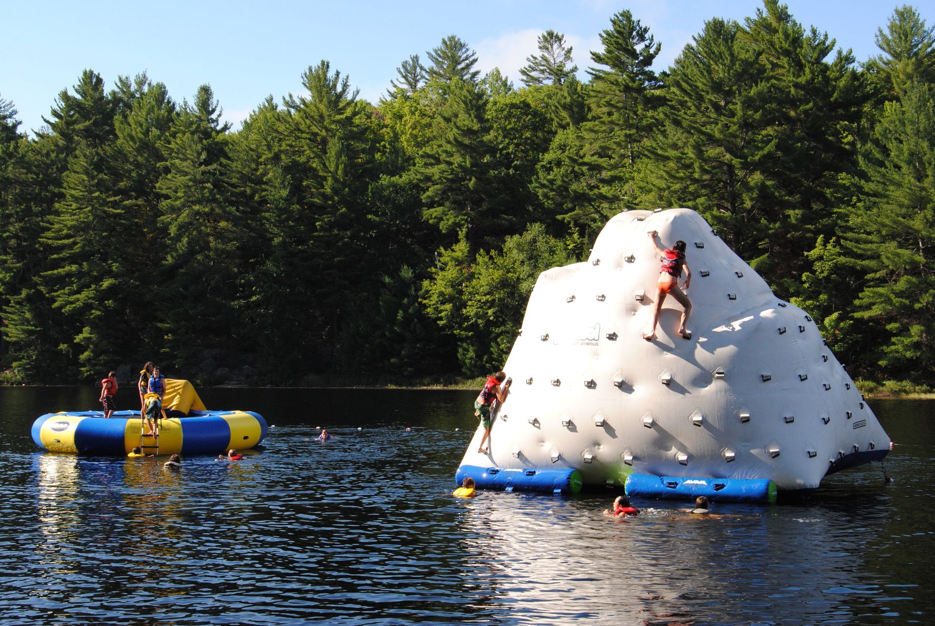 People climbing inflatable climbing wall on a lake, other playing on inflatable platforms. Trees in the background.