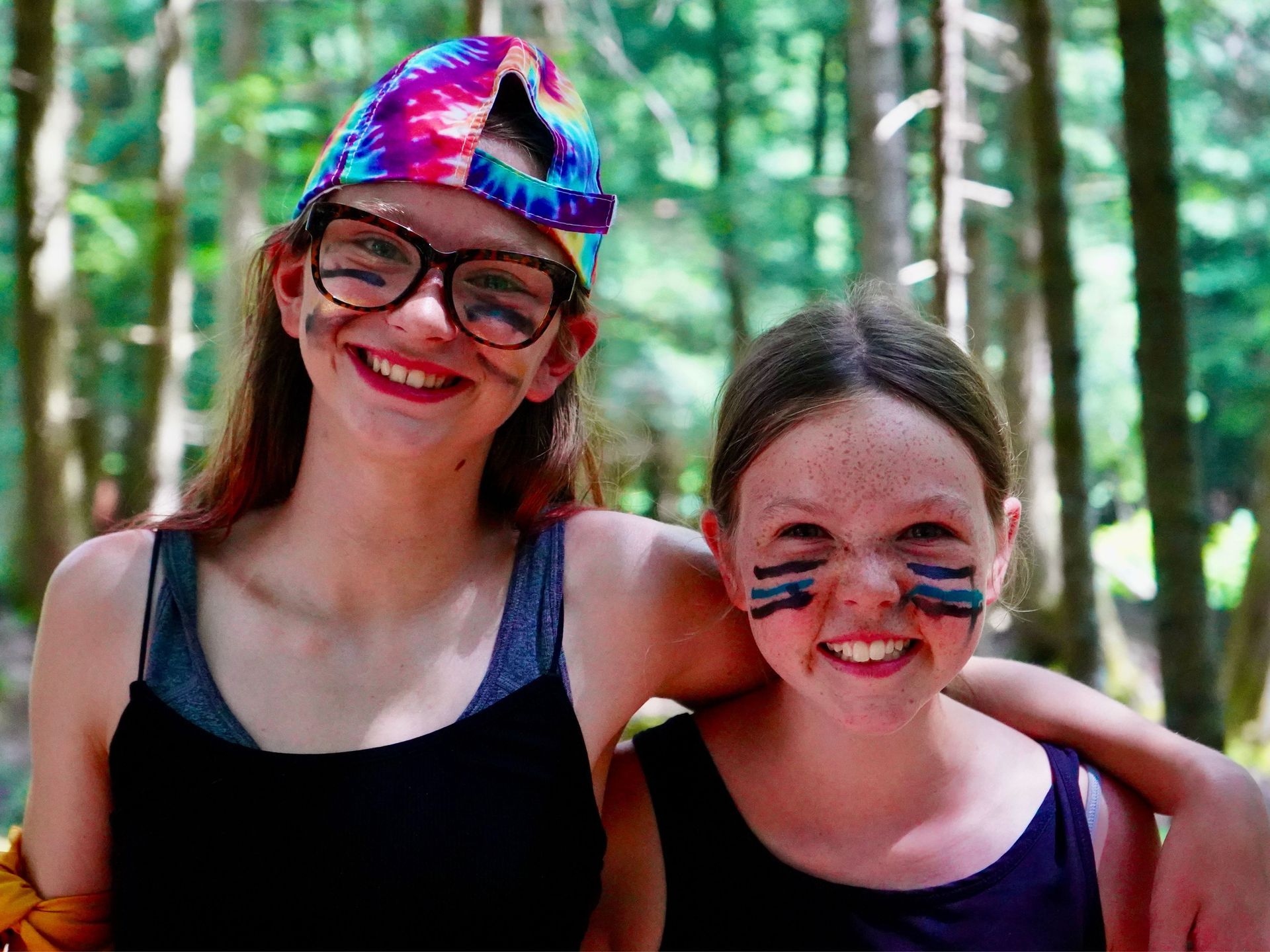 Two girls smiling, embracing, with face paint, in a wooded area. One wears glasses and a tie-dye bandana.