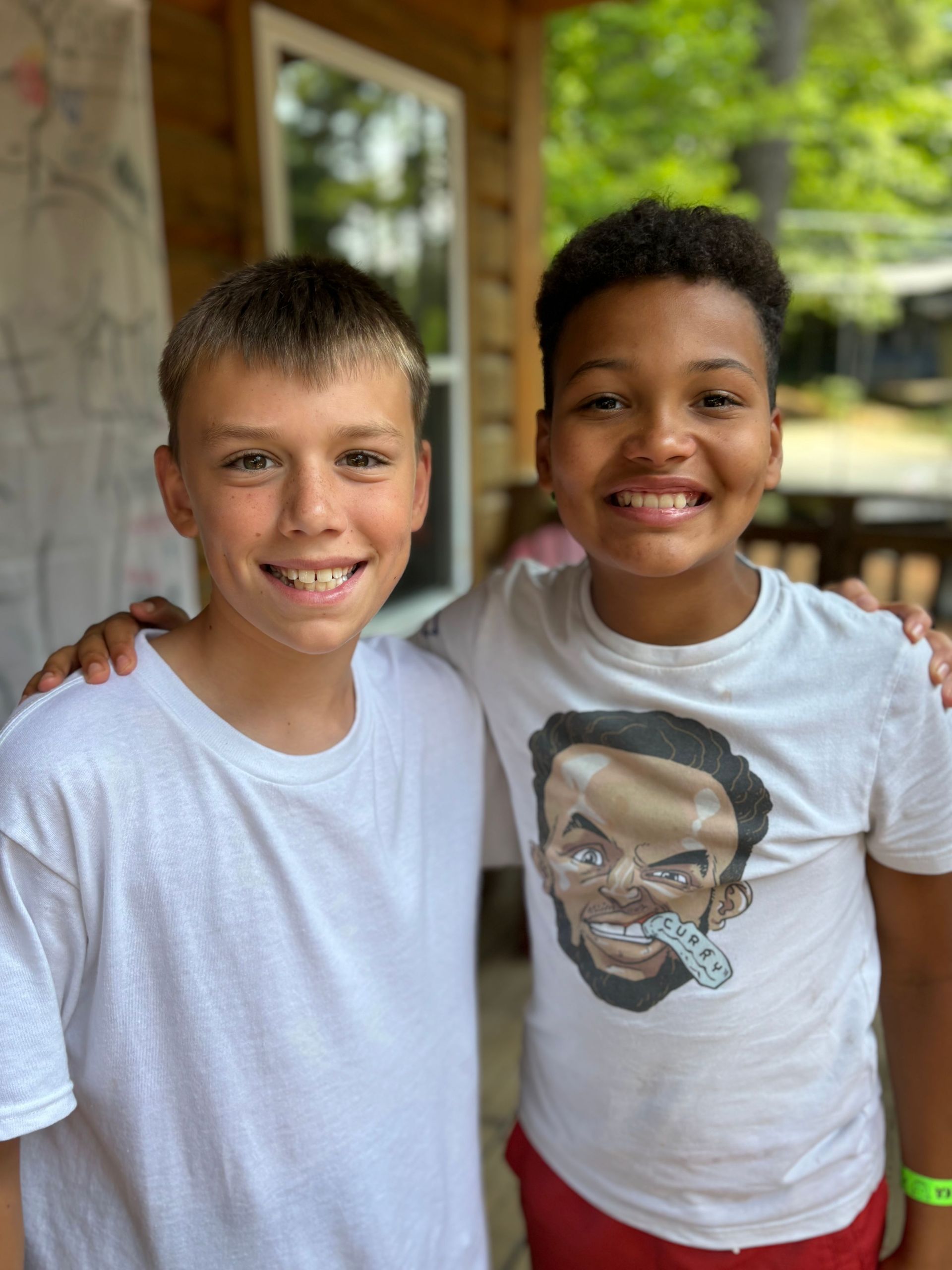 Two smiling boys with arms around each other. One boy in white t-shirt, other in graphic tee. Outside, smiling.