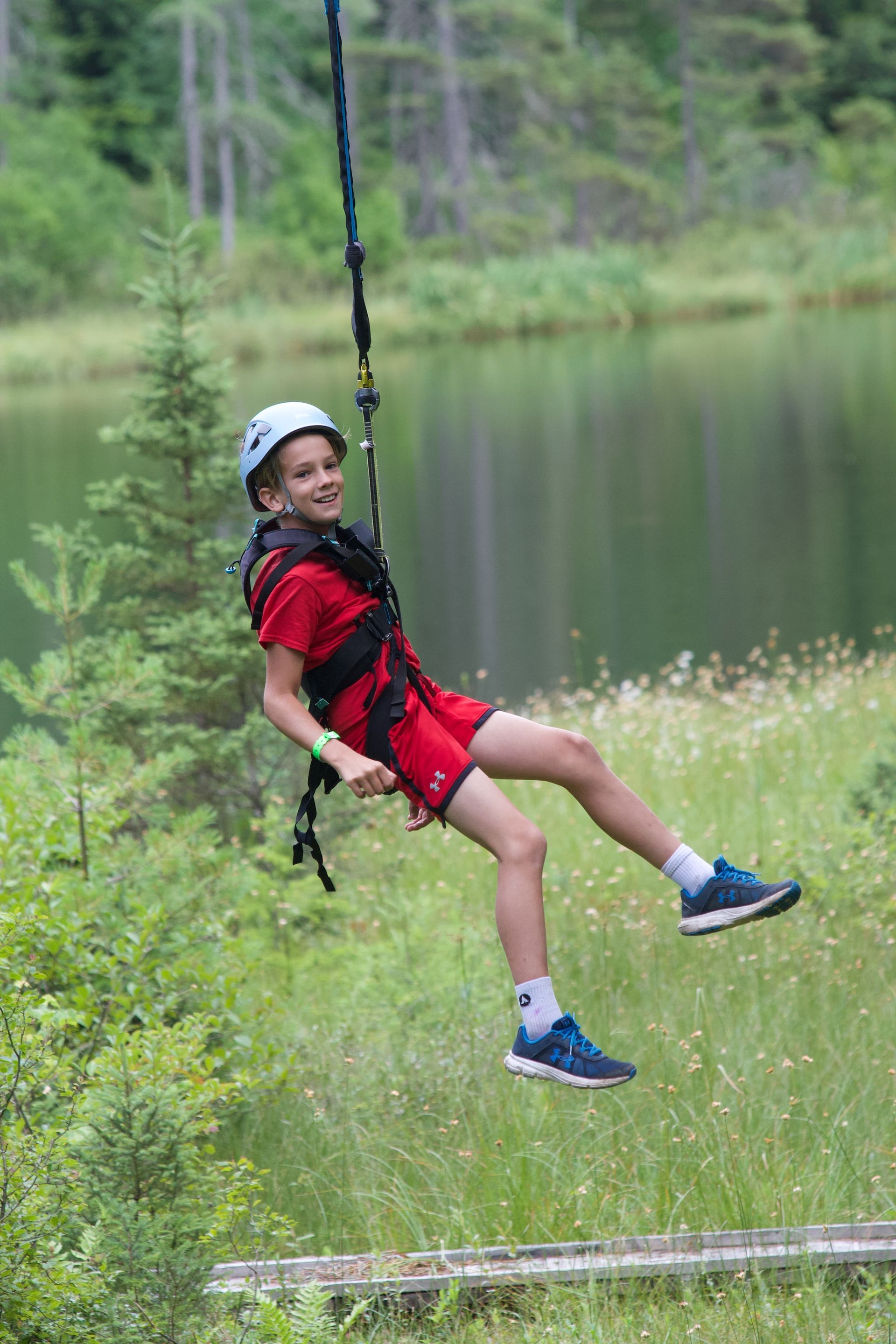 Boy in red suit and helmet suspended by bungee cord near a lake, smiling.