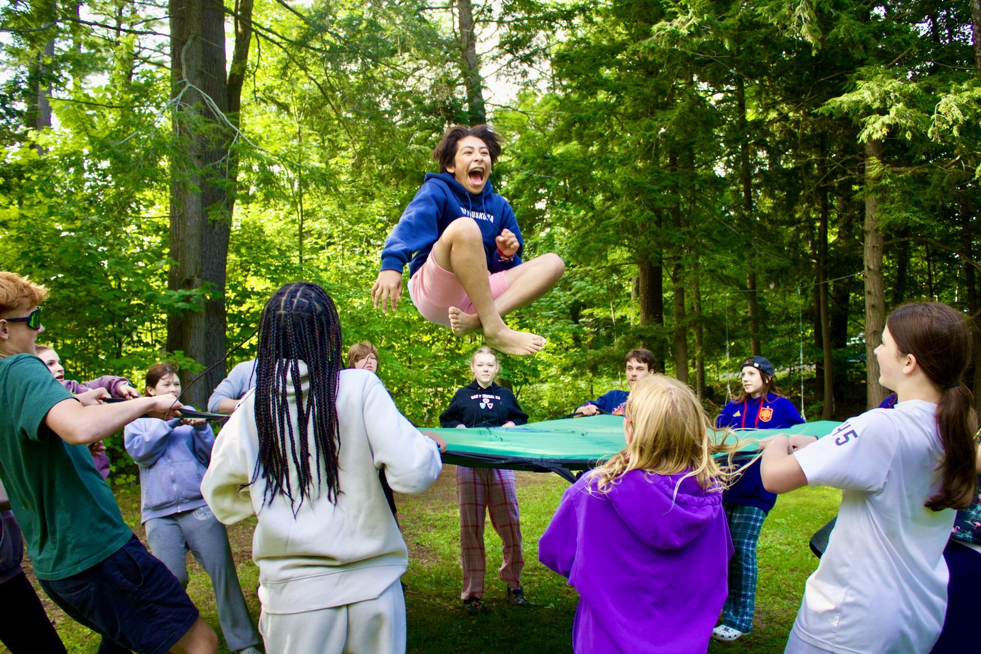 Group of people bouncing a person on a large trampoline-like net in a wooded area; person is mid-air with excited expression.