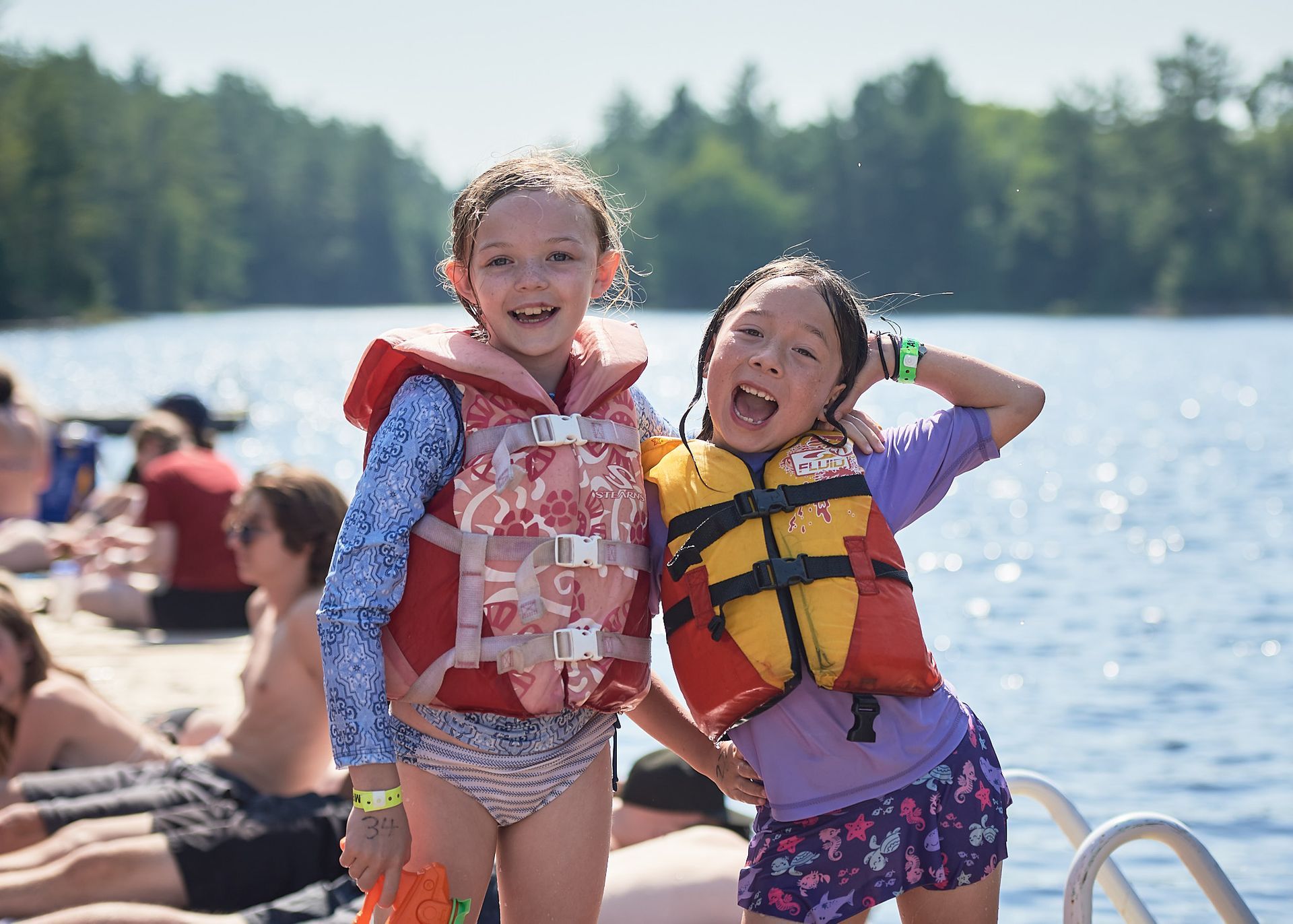 Two girls smiling by a lake, wearing life vests. Background of people on a dock, trees.