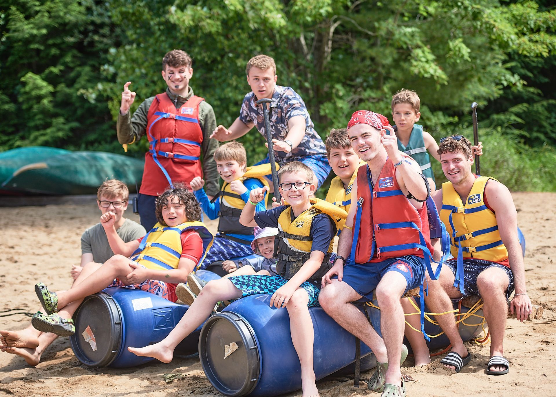 Group of people in life vests on a makeshift raft on a beach, smiling. Trees and canoes in background.