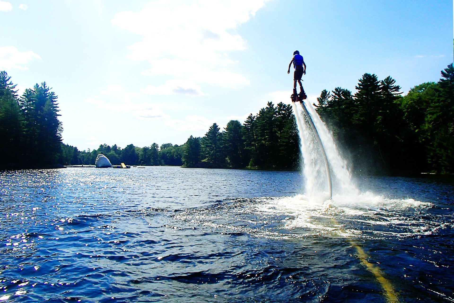 Person on a flyboard soaring over water, spraying water jets. Lake and trees in the background, blue sky.